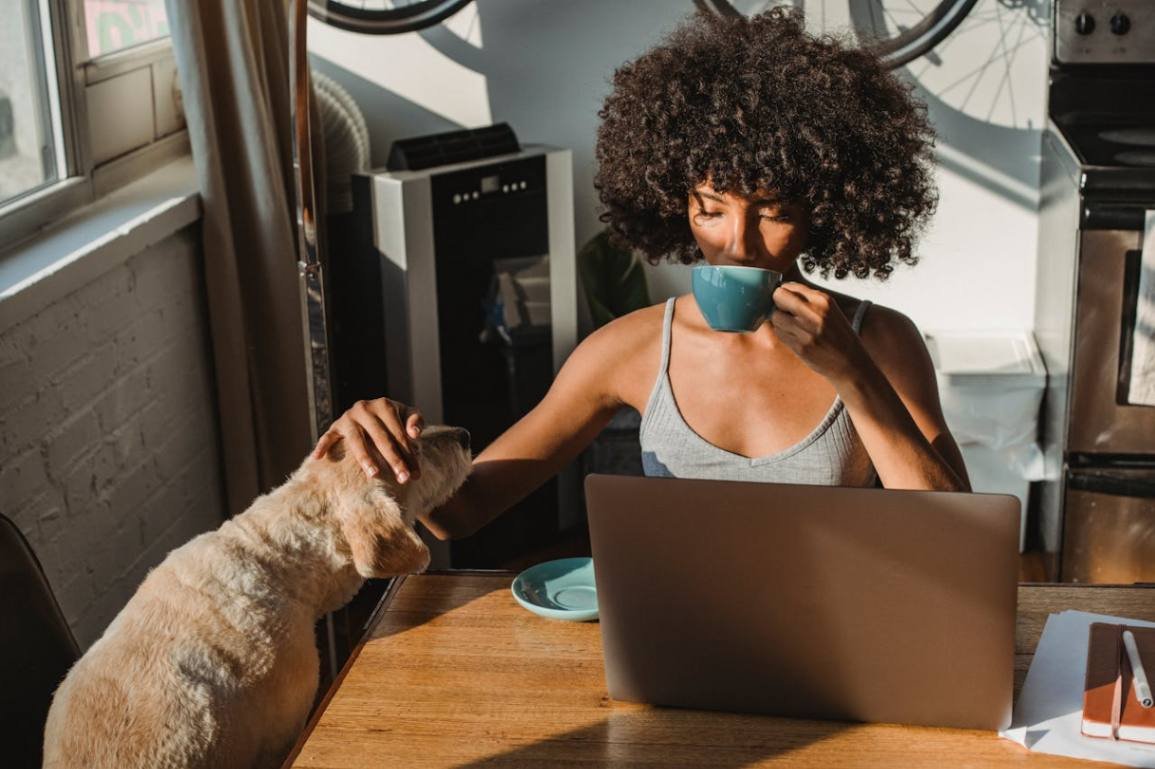 An owner and her cute dog are sitting at the table while the owner uses her laptop.